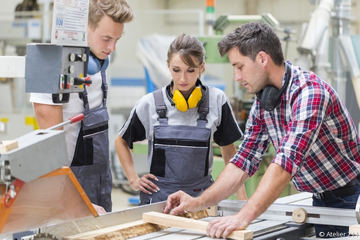 Apprentis en menuiserie autour d'un établi écoutant leur maître d'apprentissage